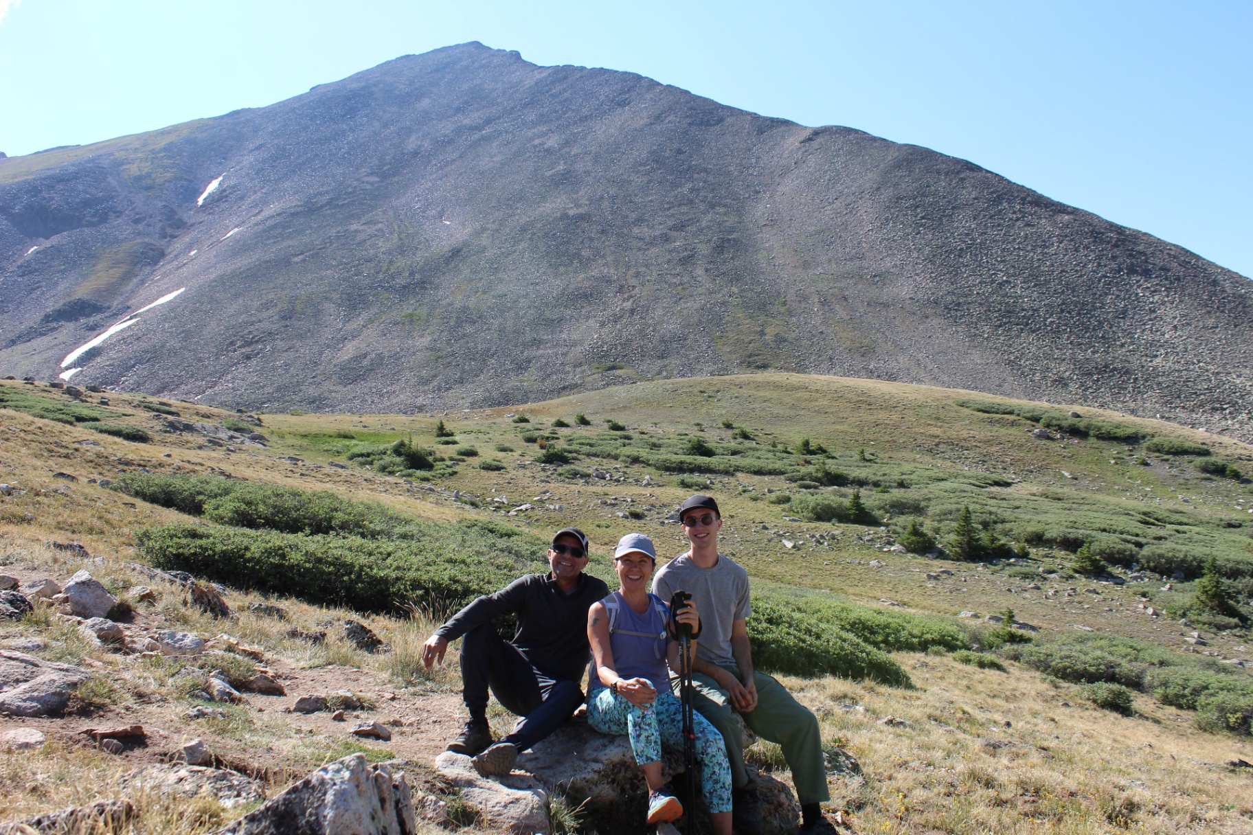 Three hikers resting on rocks in an alpine meadow below the dark scree slope of Huron Peak